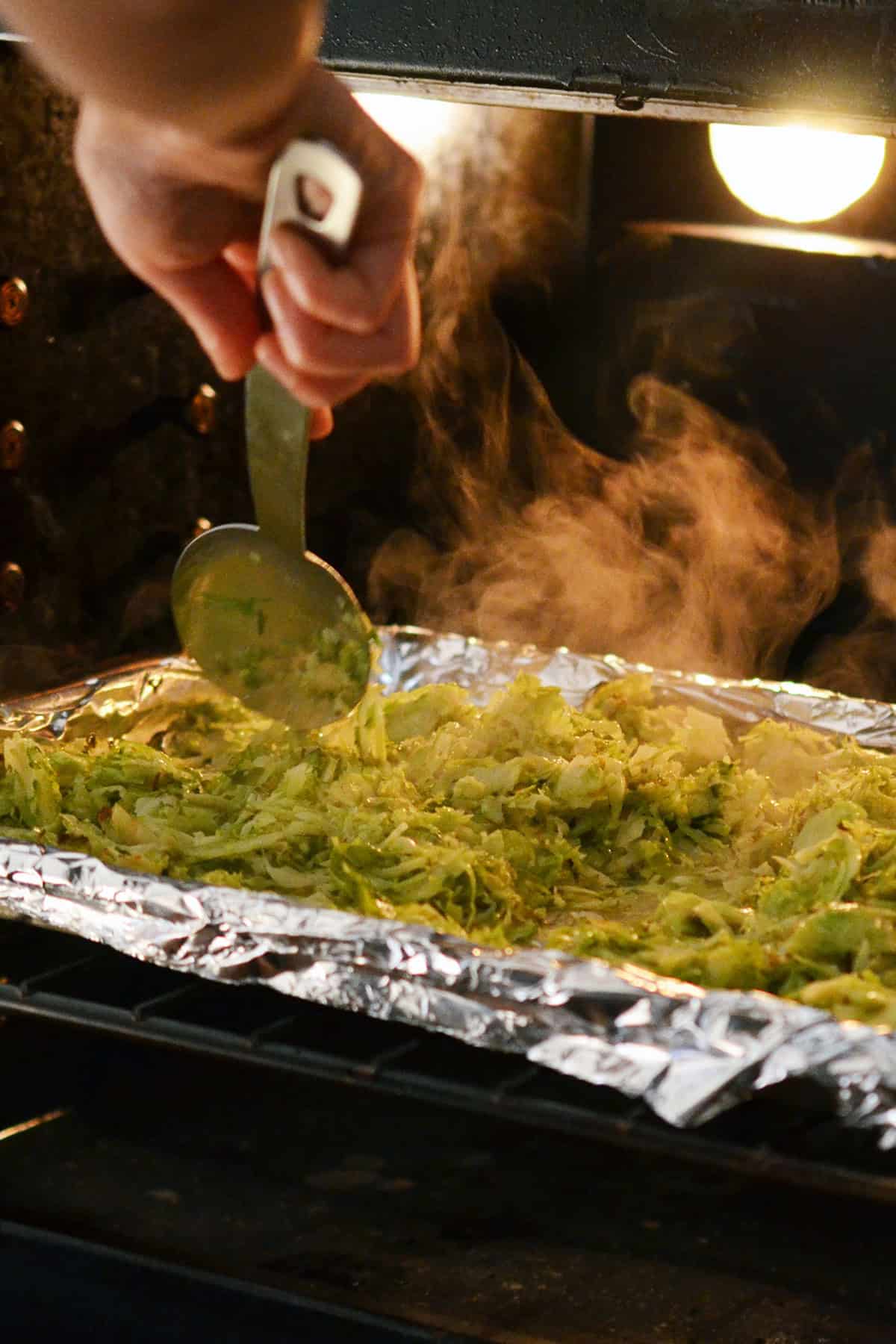 Stirring some roasted shaved Brussels sprouts in a foil lined rimmed baking sheet in the oven with a large metal spoon.