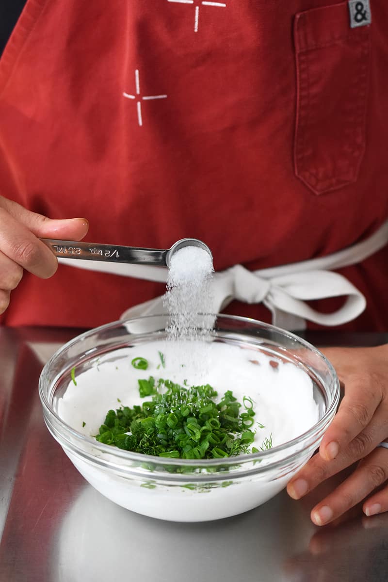 Someone in a red apron is adding salt to a small glass bowl filled with coconut yogurt and fresh green herbs.