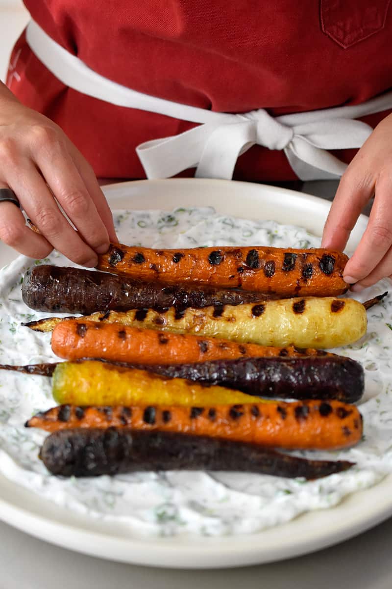 Two hands are placing grilled carrots on a platter with herbed coconut yogurt spread on the bottom.