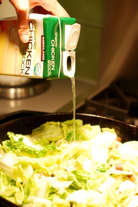 Someone is pouring chicken broth into a cast iron skillet with Napa cabbage, bacon, and onions.